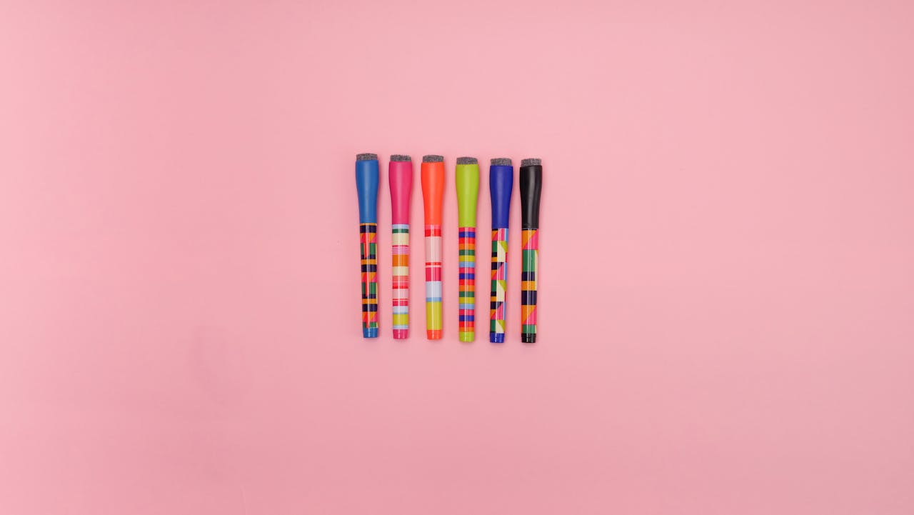 Six vibrant markers arranged on a pastel pink surface, perfect for creative and design themes.